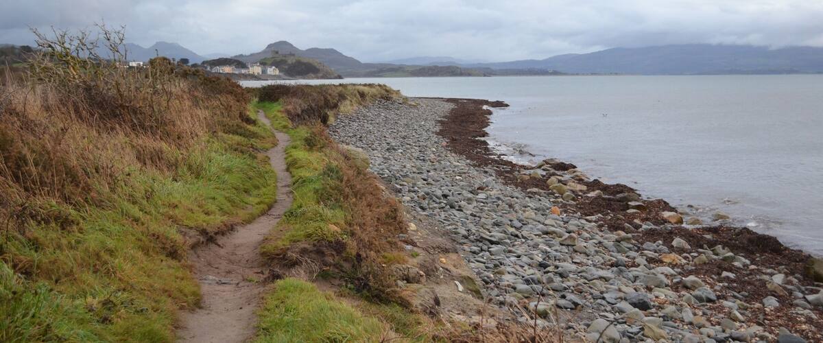 A coastal path with a rocky shoreline, a grassy trail, and distant mountains under a cloudy sky.