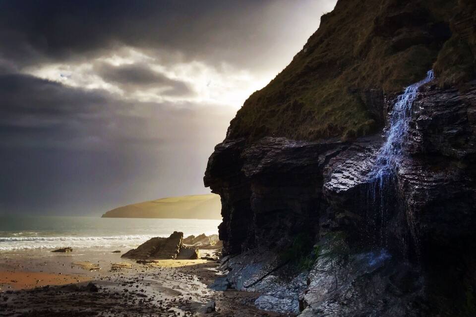 A coastal landscape with a waterfall, rocky cliffs, and a sandy beach.