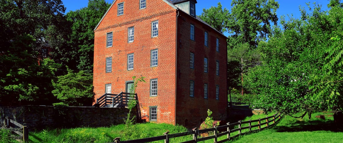A red brick building with a steep roof, surrounded by lush greenery and a wooden fence.