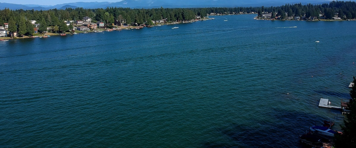 A large body of water with a forested shoreline, a small town, and snow-capped mountains in the distance.