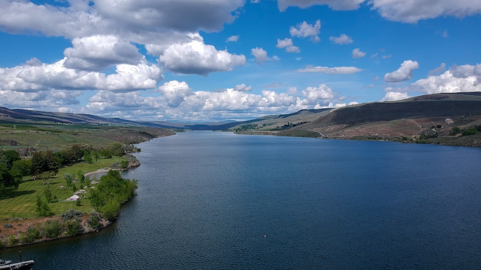 A wide river with a dock, surrounded by green hills and a clear blue sky with clouds.