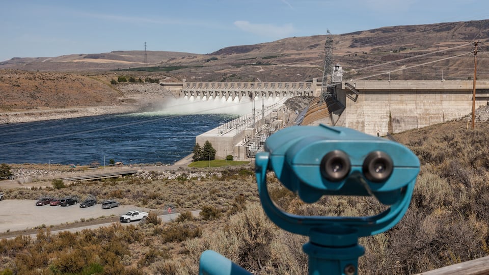 A dam with water flowing over spillways, a parking area, and a mountainous landscape.