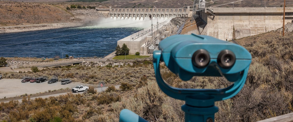 A dam with water flowing over spillways, a parking area, and a mountainous landscape.