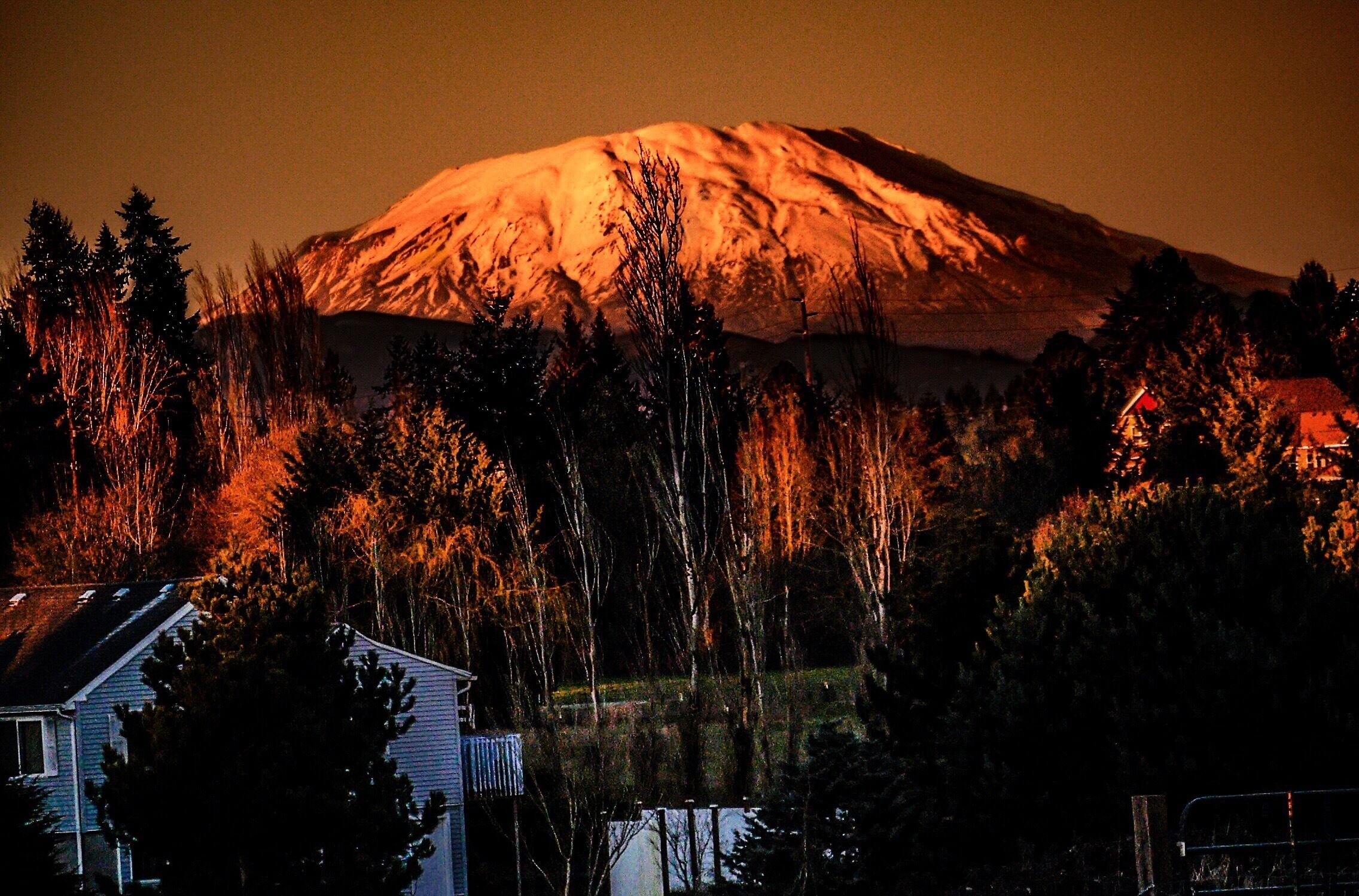 A snow-capped mountain at dusk with a house and trees in the foreground.