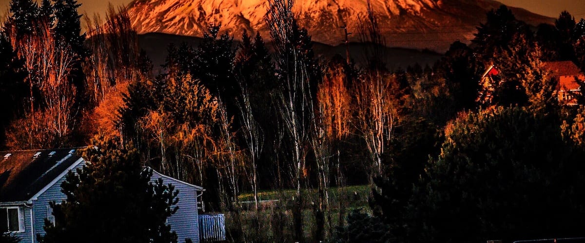 A snow-capped mountain at dusk with a house and trees in the foreground.
