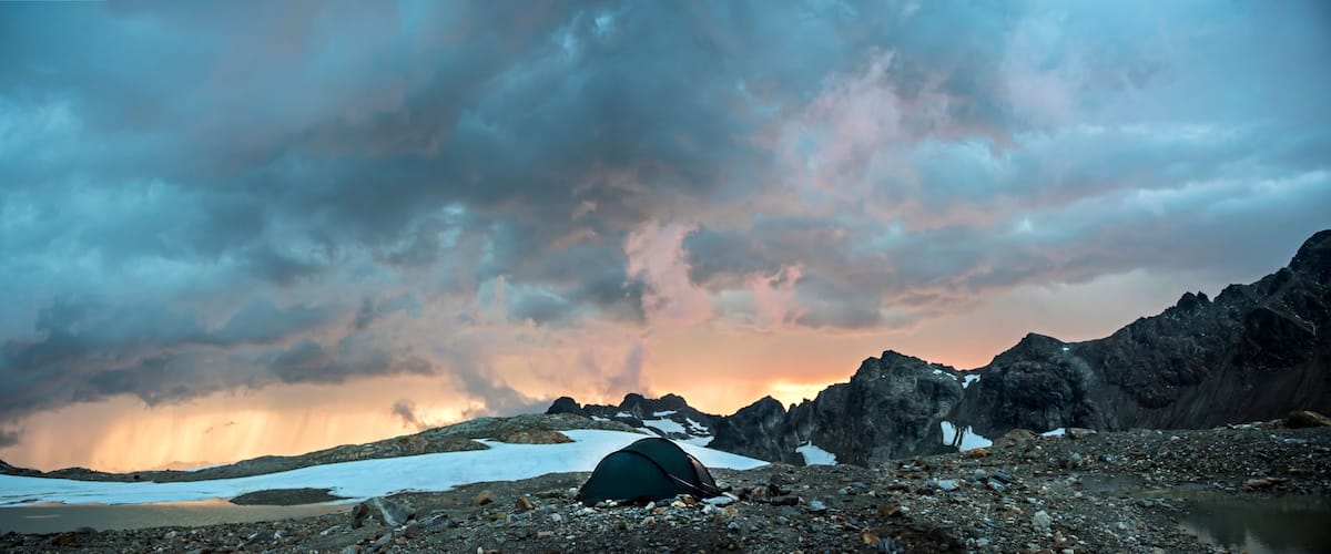 A tent on a rocky terrain with snow patches and a mountain range in the background.