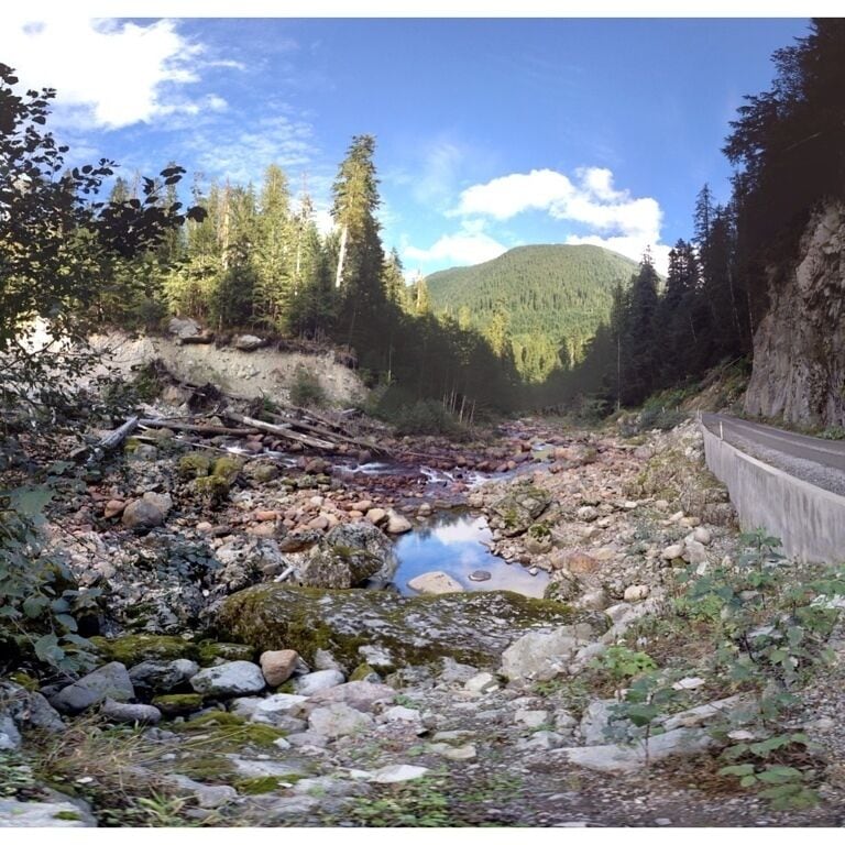 A rocky stream with a concrete road alongside, surrounded by dense forest and a mountain in the background.