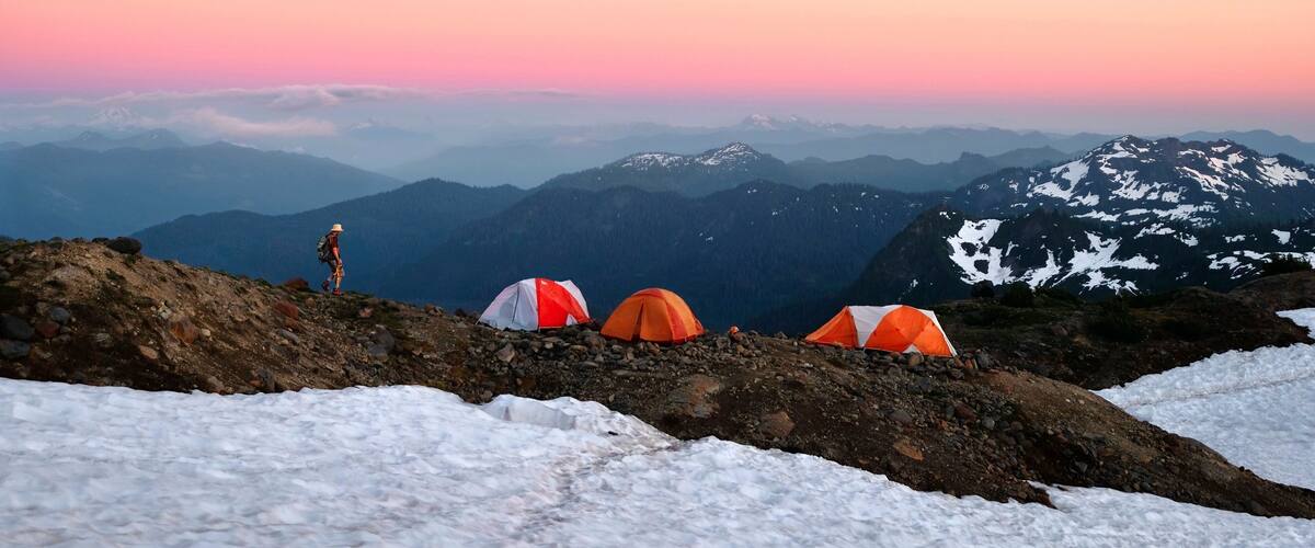 A hiker on a mountain trail with tents and snow.