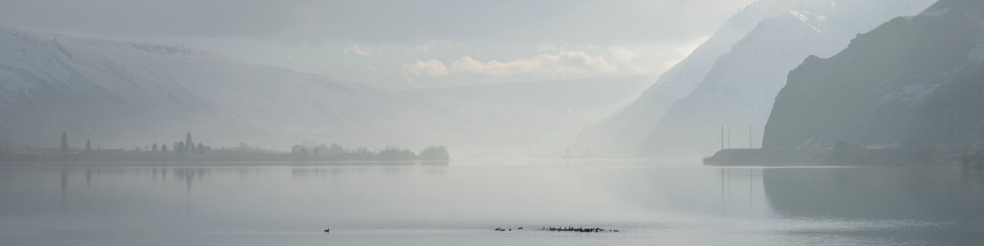 Um lago tranquilo com montanhas ao fundo e uma atmosfera nevoenta.