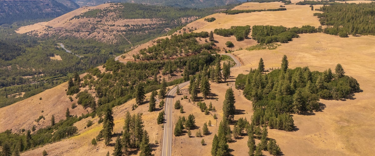 A mountain with a snow-capped peak, surrounded by forested hills and a winding road.