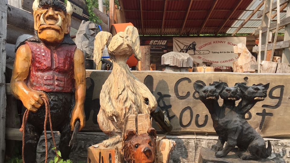 A variety of wooden carvings, including a bear, a horse, and a dog, displayed outdoors with a sign reading "CERBERUS OUTPOST."