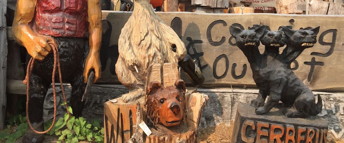 A variety of wooden carvings, including a bear, a horse, and a dog, displayed outdoors with a sign reading "CERBERUS OUTPOST."