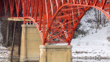 A red metal bridge over a calm river with snow-covered banks and trees in the background.