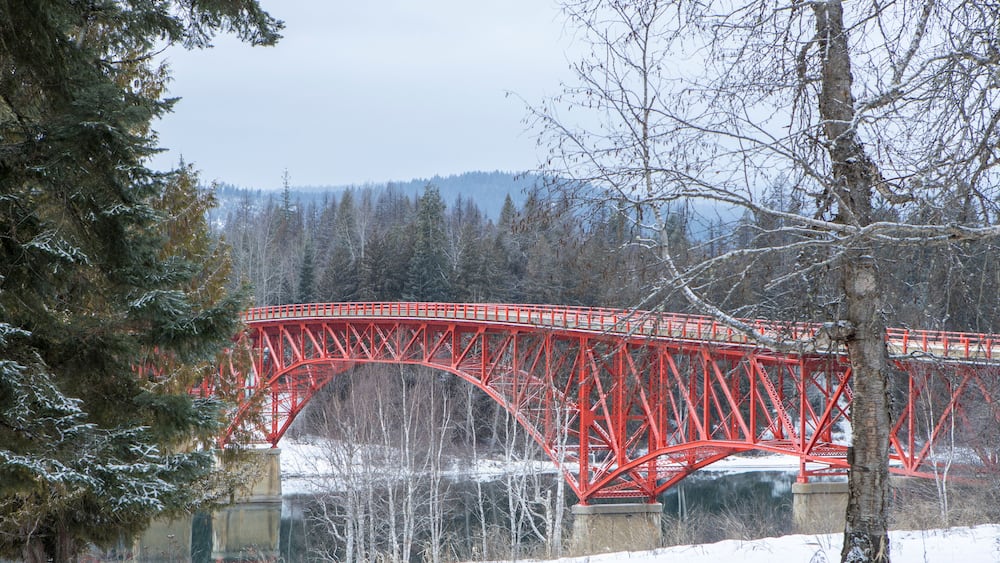 Un pont métallique rouge enjambant une rivière, entouré d’arbres et de collines couverts de neige.