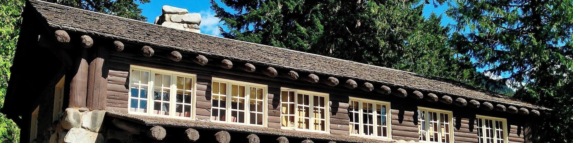 A rustic lodge with stone walls and wooden beams, surrounded by lush greenery and tall trees.