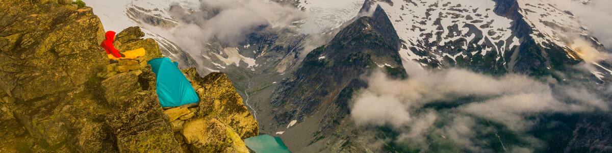 A person in red and blue gear sitting on a rocky ledge overlooking a glacier and turquoise lake.