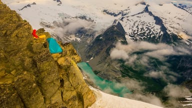 Uma pessoa vestida com roupas vermelhas e azuis sentada em um afloramento rochoso com vista para um glaciar e um lago azul-turquesa.
