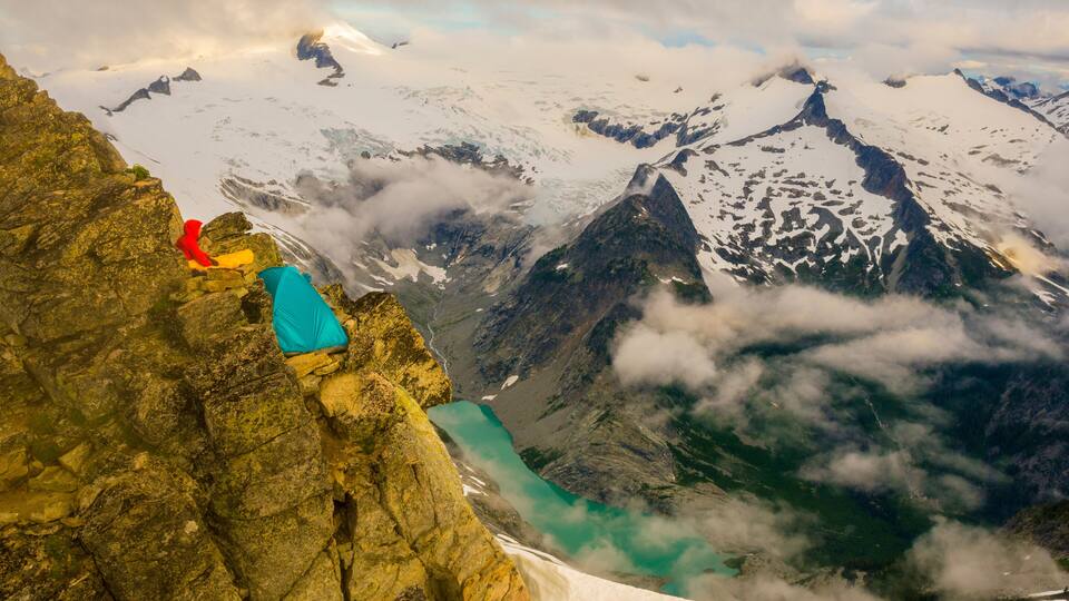 A person in red and blue gear sitting on a rocky ledge overlooking a glacier and turquoise lake.