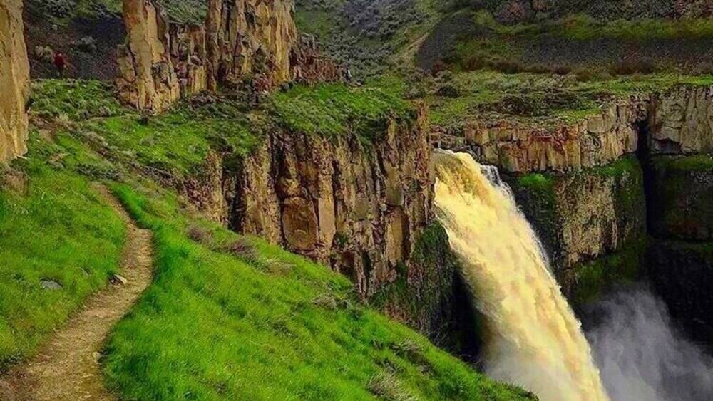 A waterfall cascading down a rocky cliff, surrounded by lush green hills and a clear sky.