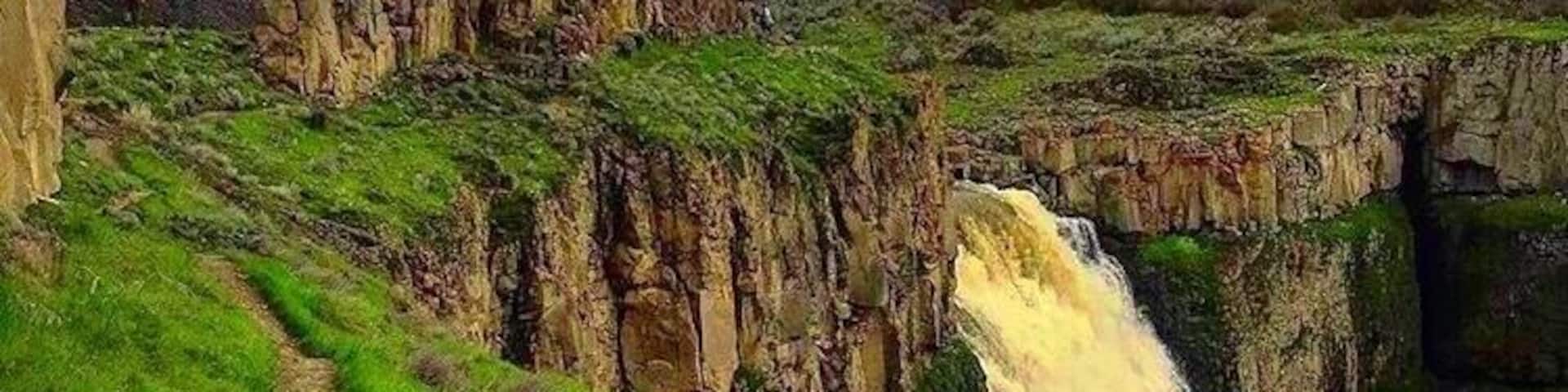 A waterfall cascading down a rocky cliff, surrounded by lush green hills and a clear sky.