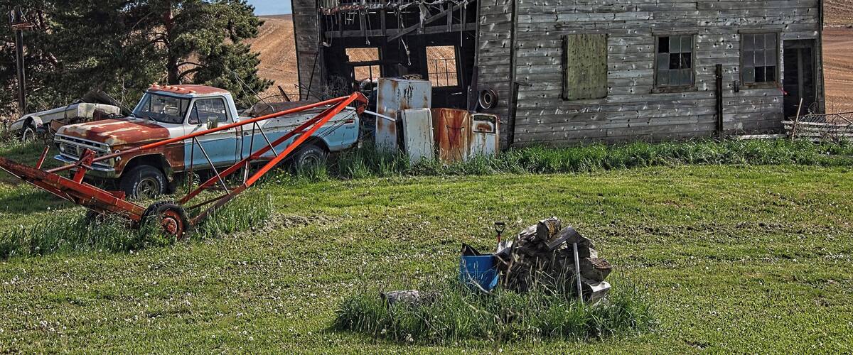 An abandoned wooden barn with a rusted roof, surrounded by overgrown grass and a vintage truck.