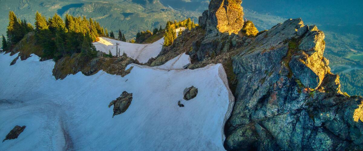 A mountain landscape with snow-covered peaks, rocky outcrops, and dense forests.