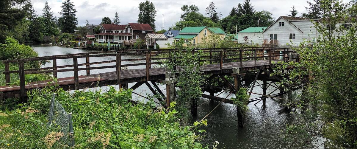 A wooden bridge over a river with houses on the opposite bank.