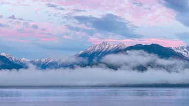 A mountain range with snow-capped peaks, a lake in the foreground, and a sky with pink and blue hues.
