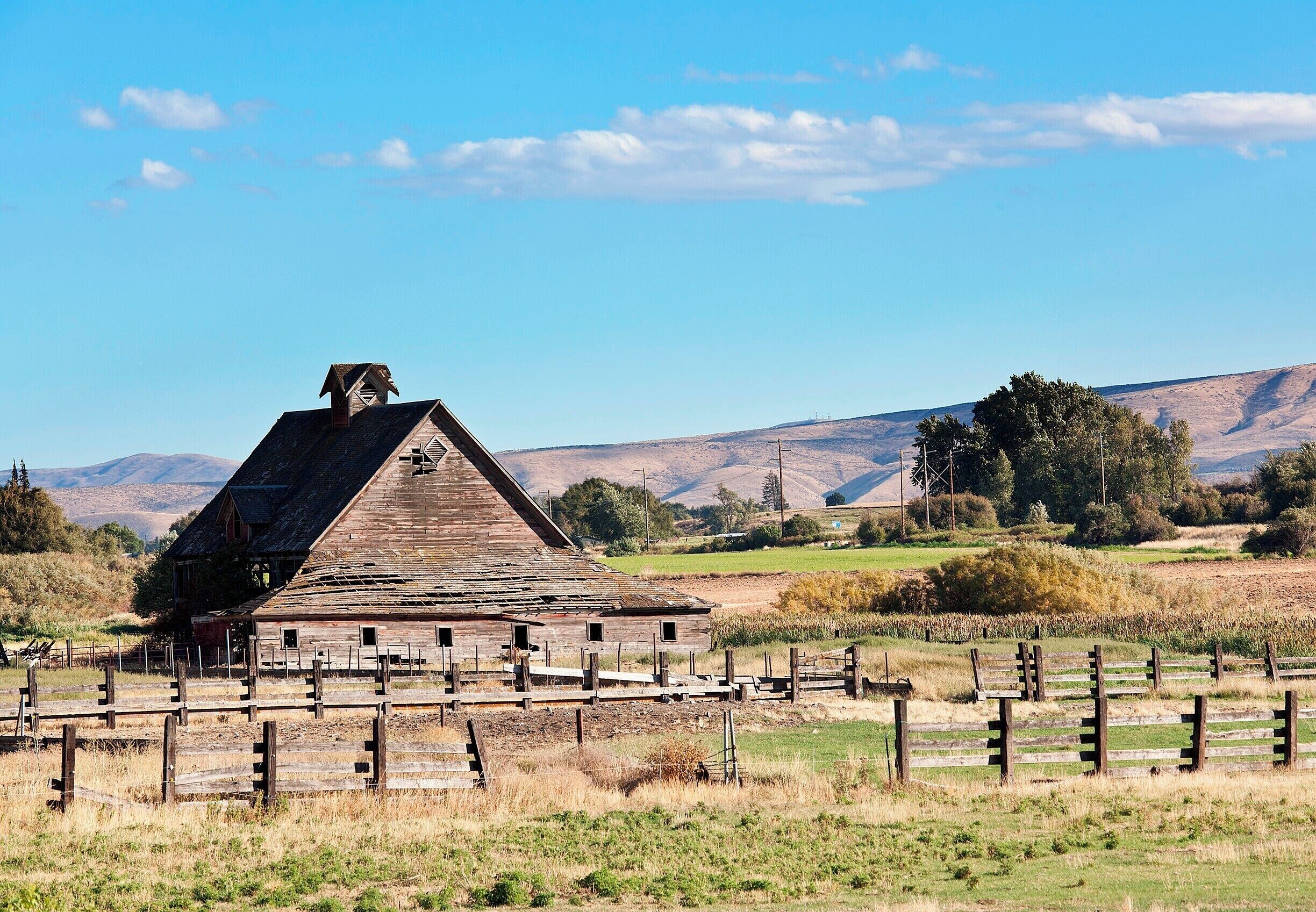A rustic barn with a steep roof, surrounded by a wooden fence in a rural landscape with rolling hills and sparse vegetation.