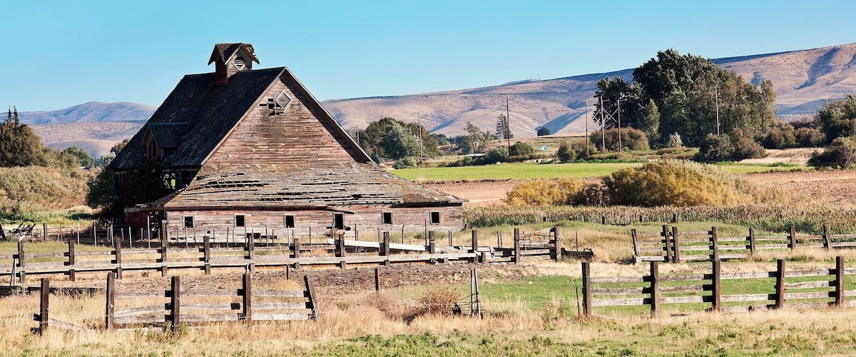 A rustic barn with a steep roof, surrounded by a wooden fence in a rural landscape with rolling hills and sparse vegetation.