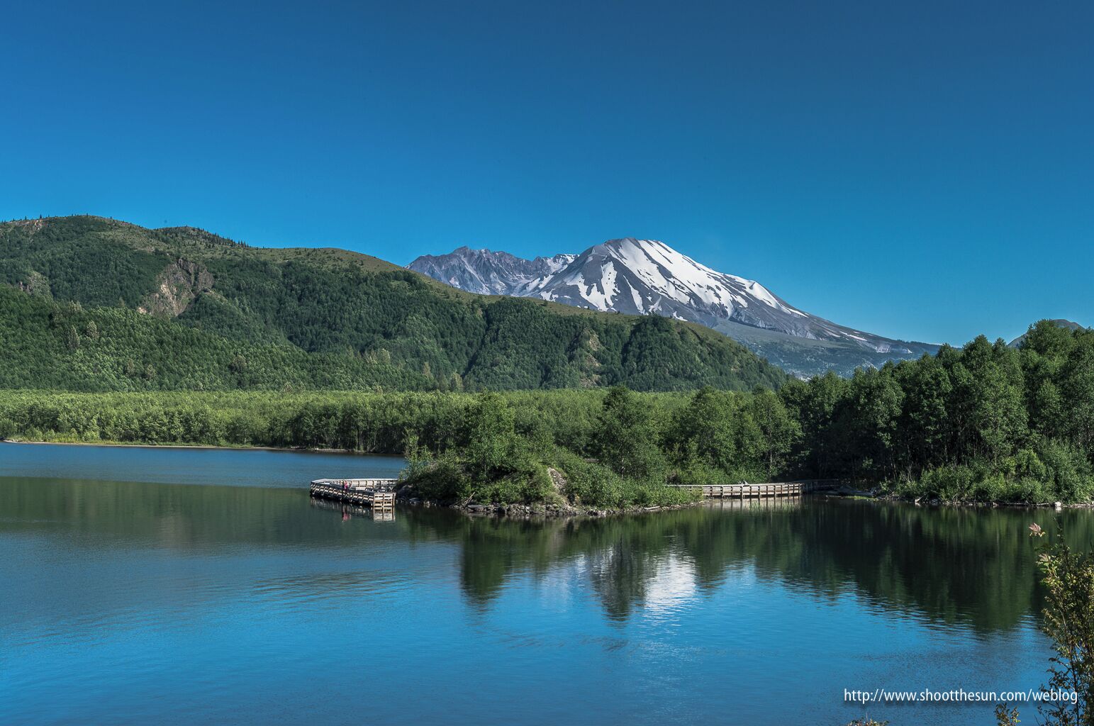 A lake with a wooden dock, surrounded by dense green forest and a snow-capped mountain in the background.