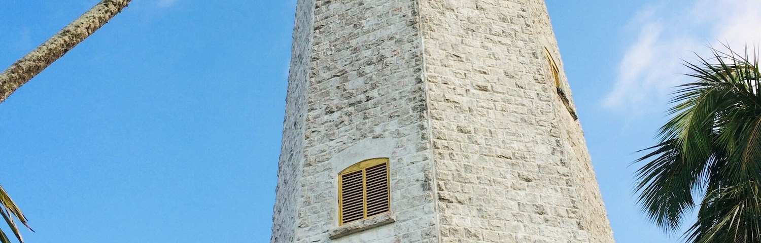 Un alto faro de piedra blanca con dos ventanas, rodeado de palmeras y un cielo azul despejado.