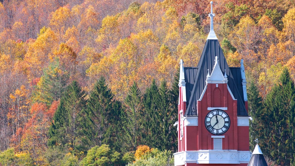 A red church with a clock tower and a forest in autumn colors in the background.