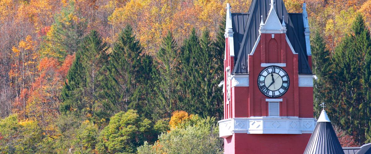 A red church with a clock tower and a forest in autumn colors in the background.