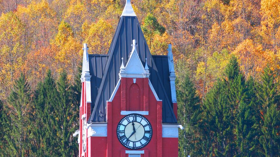 A red church with a clock tower and a steeple, surrounded by autumn-colored trees.