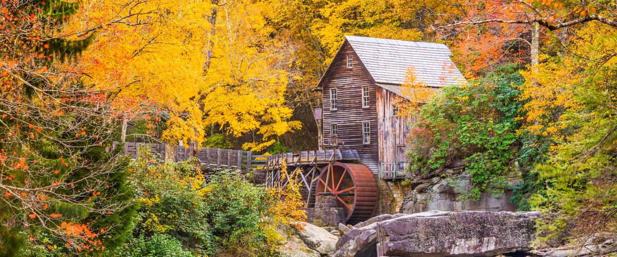 A wooden mill with a water wheel, surrounded by autumn foliage and a cascading waterfall.