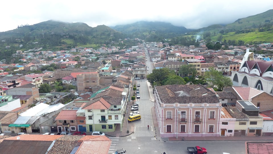 A town with a central street flanked by buildings, a church with a distinctive roof, and mountains in the background.