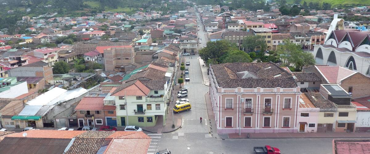 A town with a central street flanked by buildings, a church with a distinctive roof, and mountains in the background.