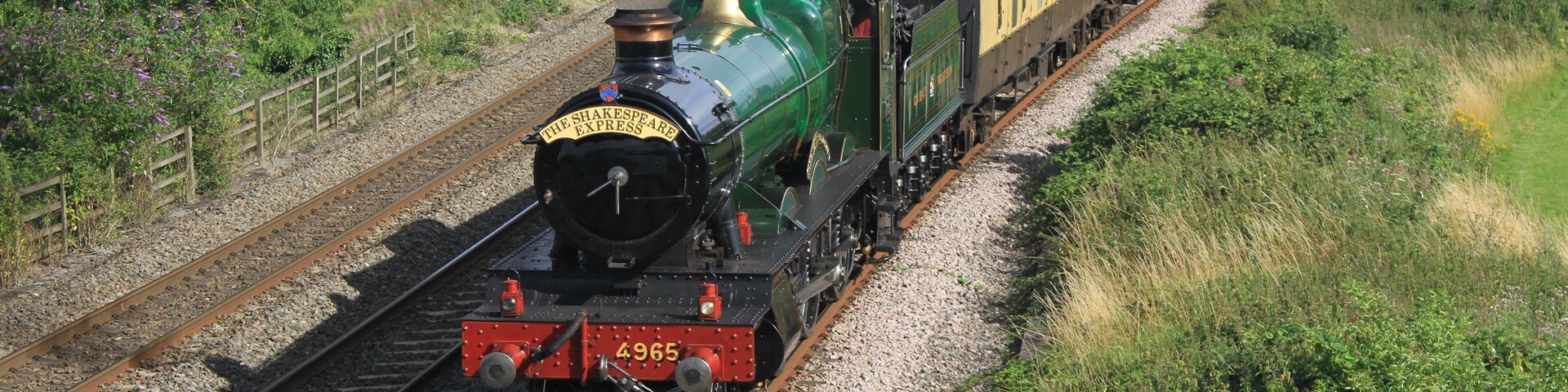 Une locomotive à vapeur verte tirant des voitures de voyageurs sur une voie ferrée, le tout étant entouré de verdure.