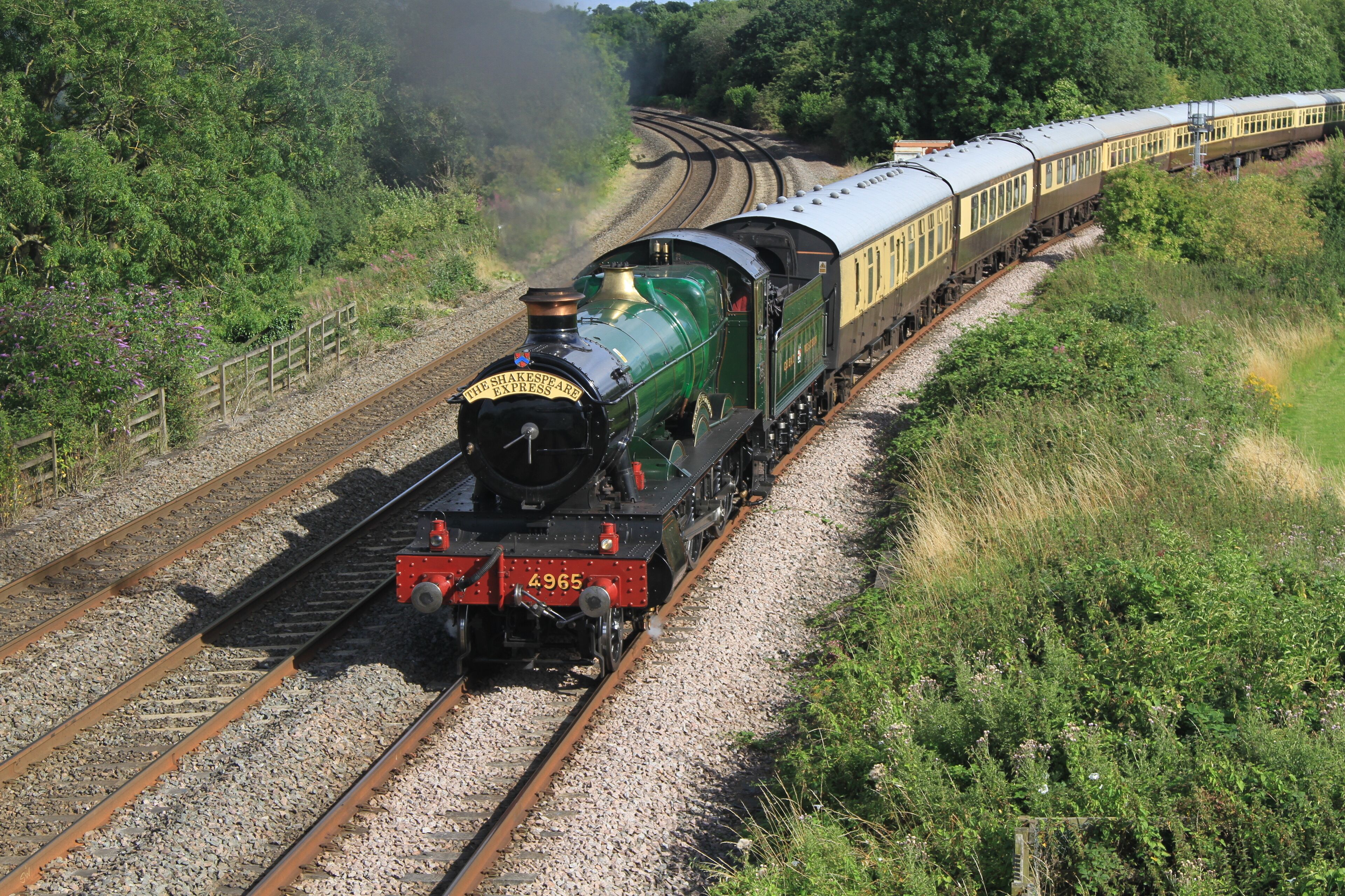 A green steam locomotive pulling passenger cars on a railway track surrounded by greenery.