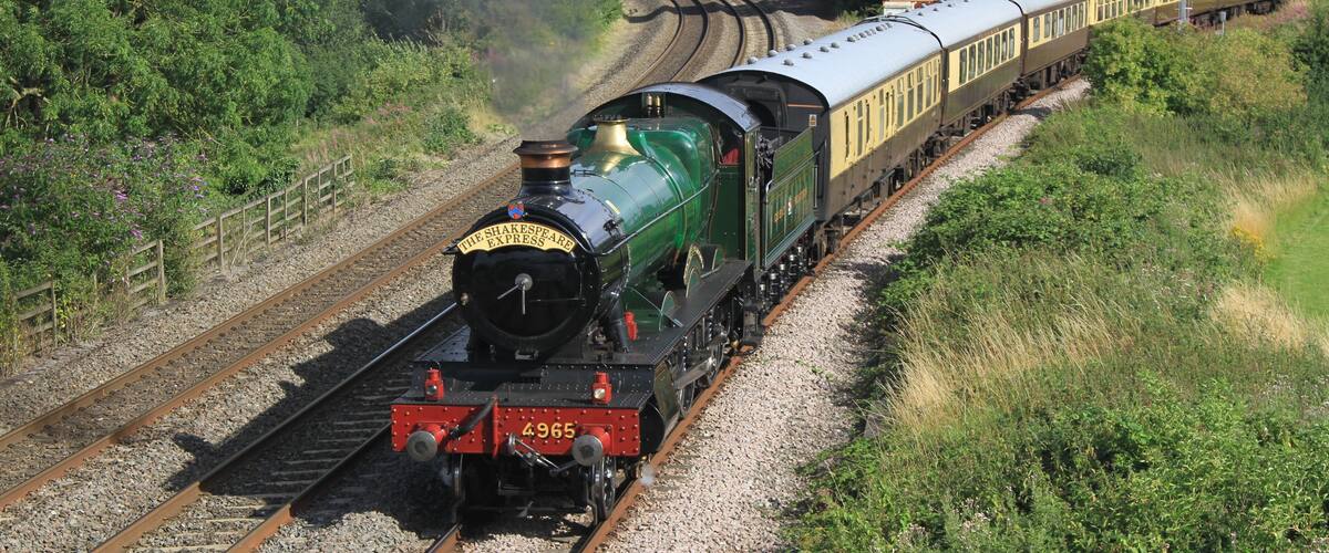 A green steam locomotive pulling passenger cars on a railway track surrounded by greenery.