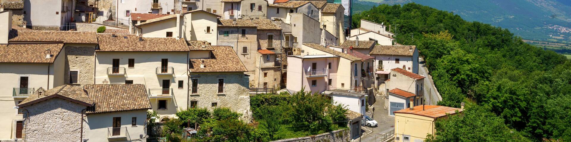 A historic town with terracotta-roofed buildings, a prominent church tower, and a mountain range in the background.
