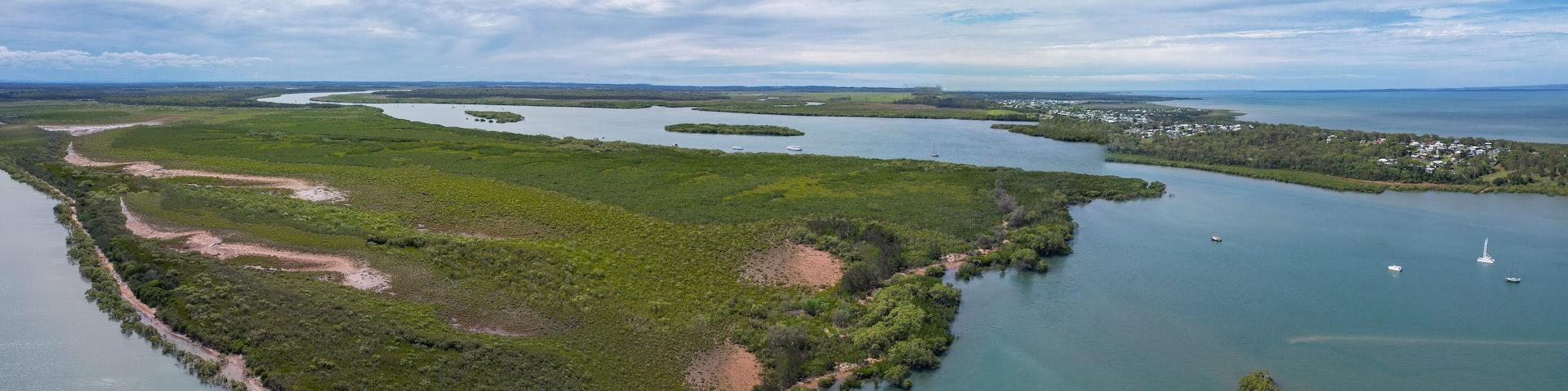 Aerial view of the serpentine river snaking through verdant mangroves, meeting the cerulean sea under a cloudy sky, River Heads, Queensland, Australia.