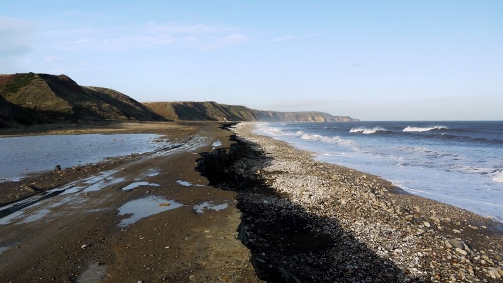 A rocky beach with waves crashing onto the shore, cliffs in the background, and a clear sky.