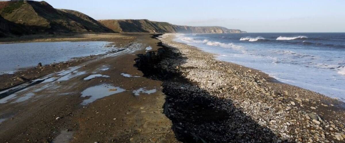 A rocky beach with waves crashing onto the shore, cliffs in the background, and a clear sky.