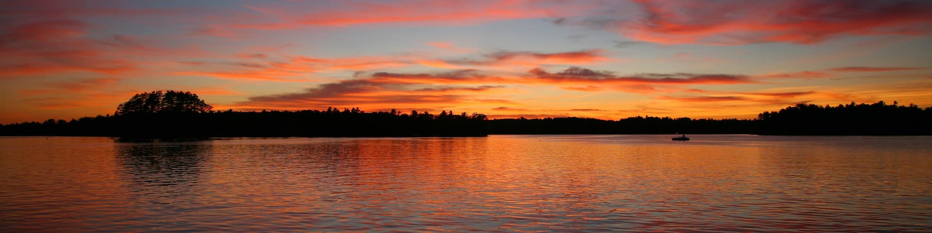 A sunset over a calm lake with a silhouette of a tree-covered island and a small boat.