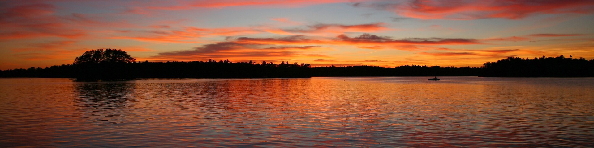 A sunset over a calm lake with a silhouette of a tree-covered island and a small boat.