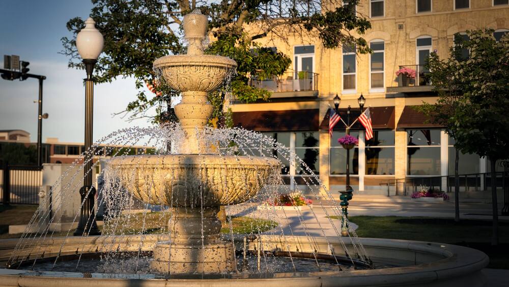 A multi-tiered stone fountain with water jets, surrounded by a paved area with a building in the background.