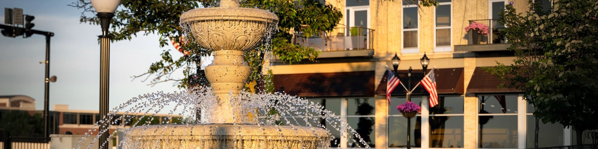 A multi-tiered stone fountain with water jets, surrounded by a paved area with a building in the background.