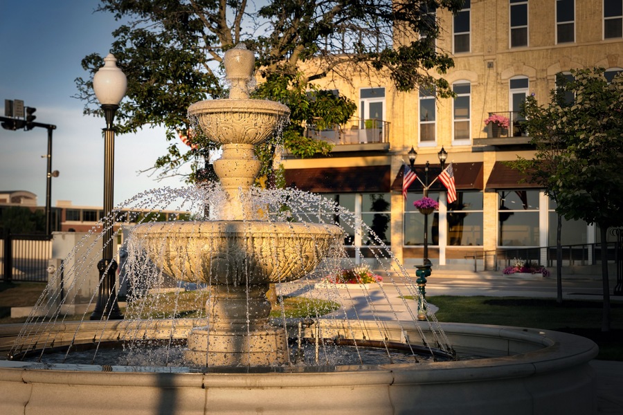 A multi-tiered stone fountain with water jets, surrounded by a paved area with a building in the background.